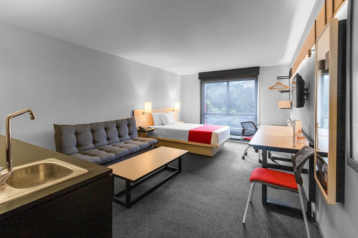 A compact hotel room with a sofa, a bed, a desk, and a kitchenette.