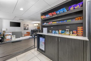 A snack kiosk with shelves stocked with various chips and snacks, a refrigerator with beverages, and a counter with magazines and newspapers.
