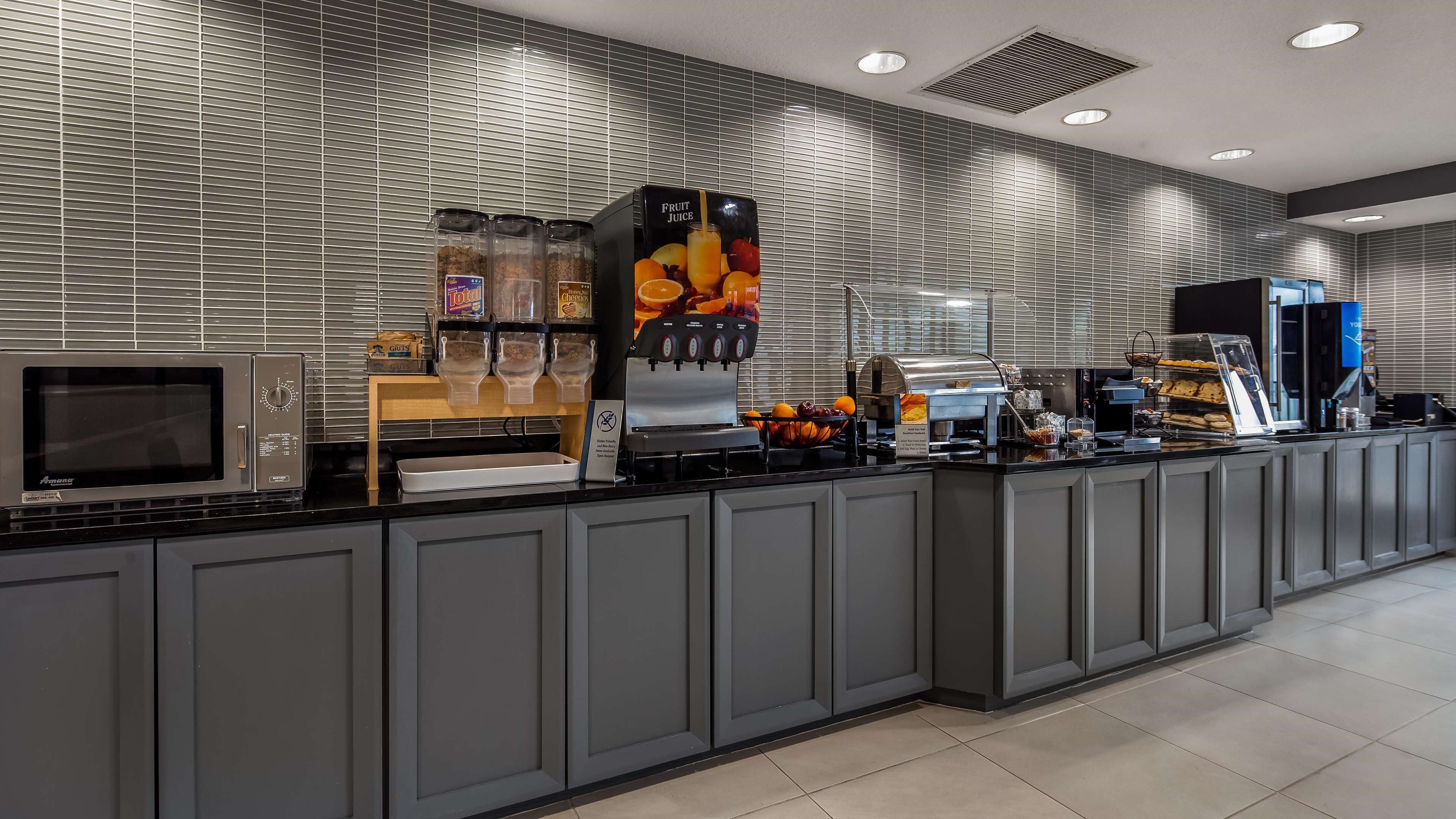 A modern self-service breakfast area with a microwave, beverage dispensers, and a variety of food options.