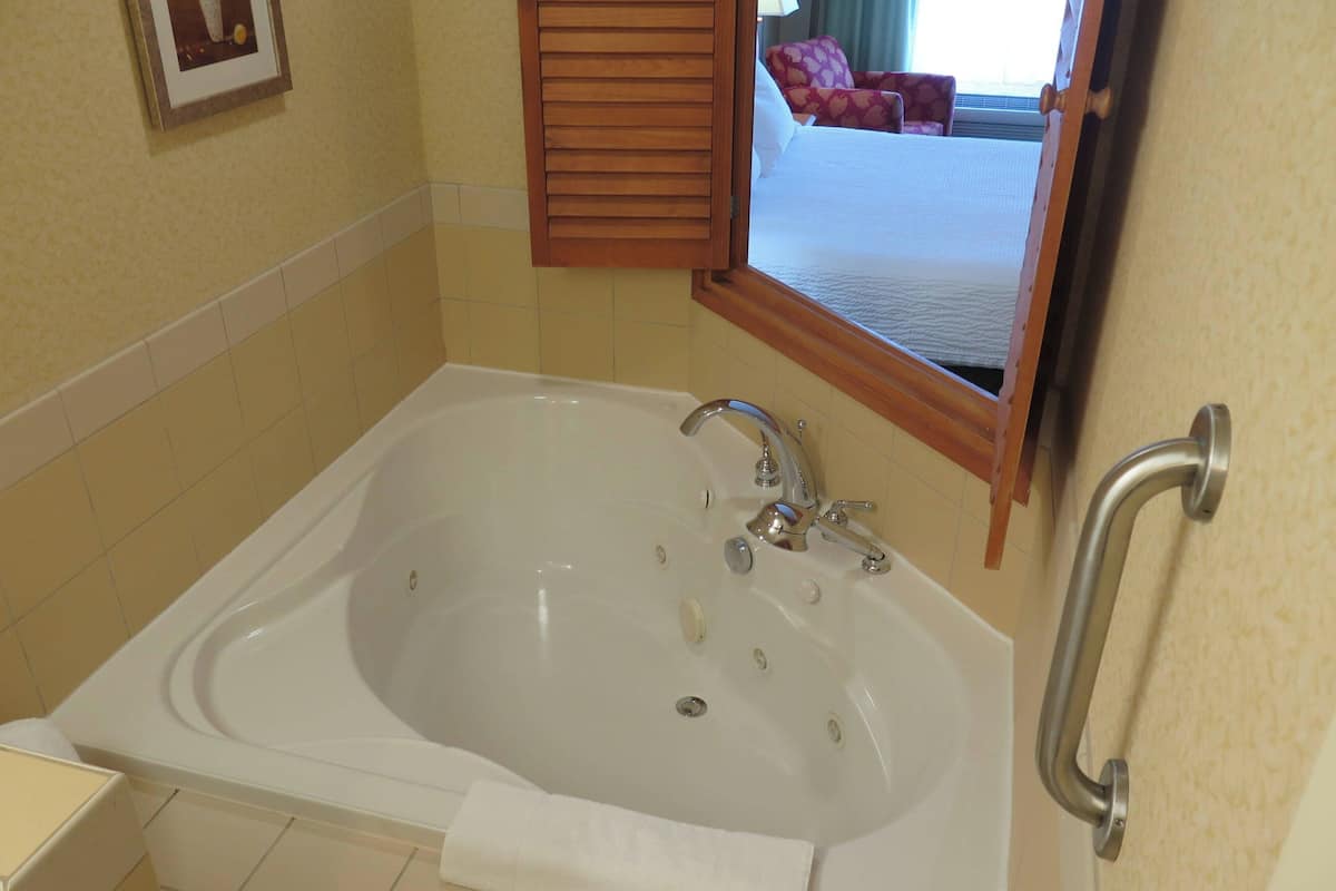 Een badkamer met een groot bad, een handgreep en een spiegel die een bed en een raam weerspiegelt.
