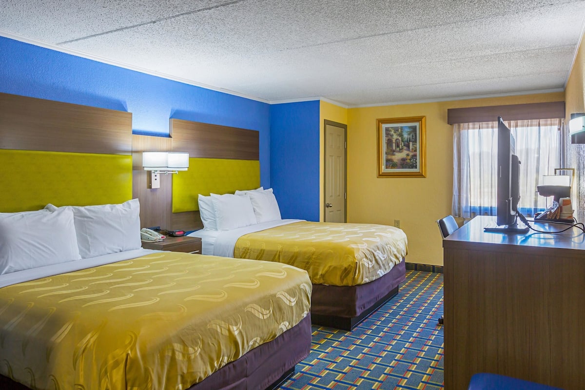 A hotel room with two beds, a desk, a chair, a nightstand, and a painting on the wall.