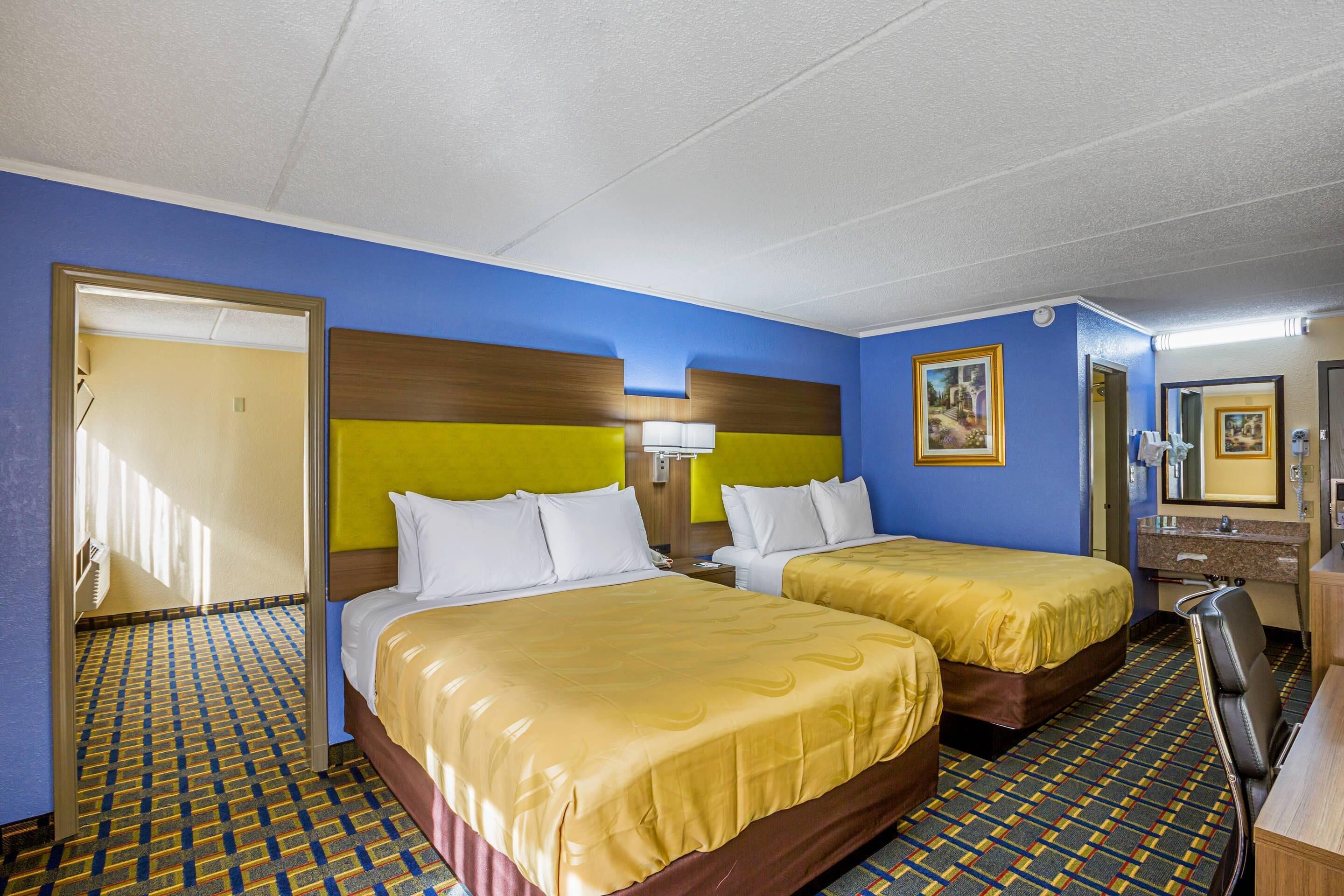Habitación de hotel con dos camas, un escritorio, una silla, un espejo y un baño con lavabo y espejo.