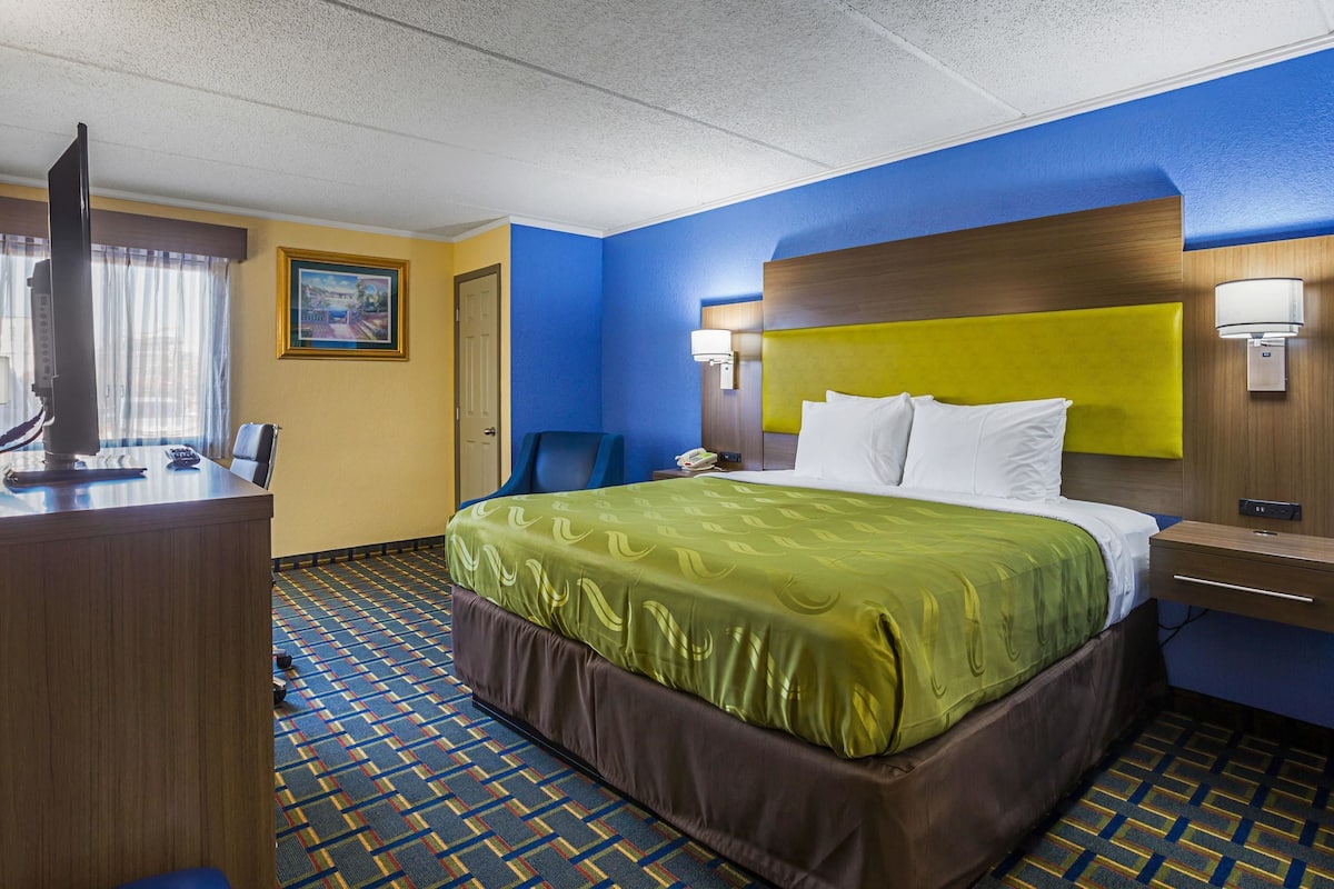 A hotel room with a bed, a desk, a chair, a television, and a painting on the wall.