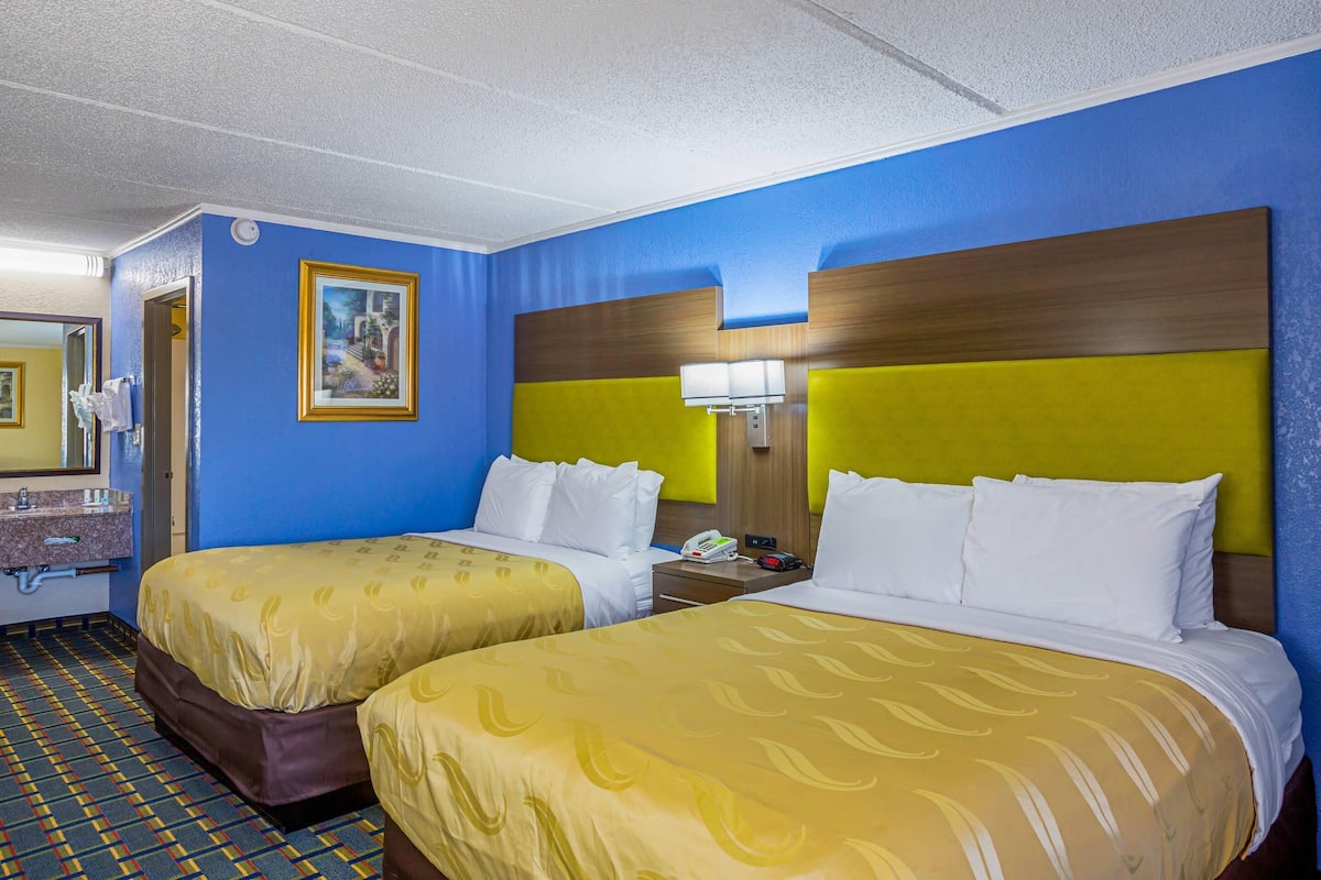 A hotel room with two beds, a bathroom with a sink and mirror, and a painting on the wall.