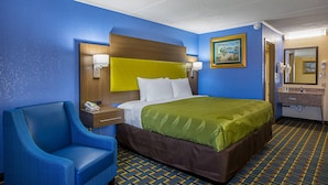A hotel room with a blue and yellow color scheme, a bed with a green bedspread, a blue armchair, a bedside table with a lamp, and a bathroom with a mirror and sink. - Quality Inn & Suites near Six Flags East (Atlanta)