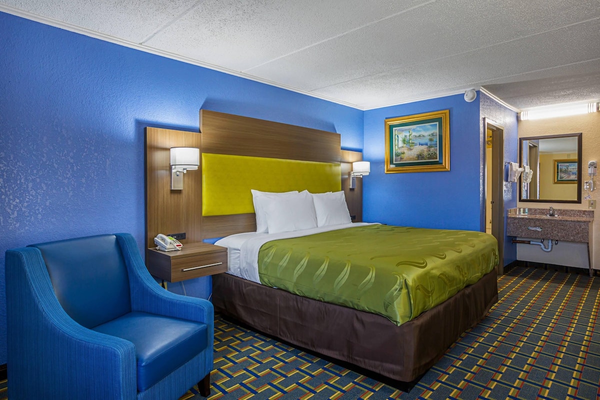 A hotel room with a blue and yellow color scheme, a bed with a green bedspread, a blue armchair, a bedside table with a lamp, and a bathroom with a mirror and sink.