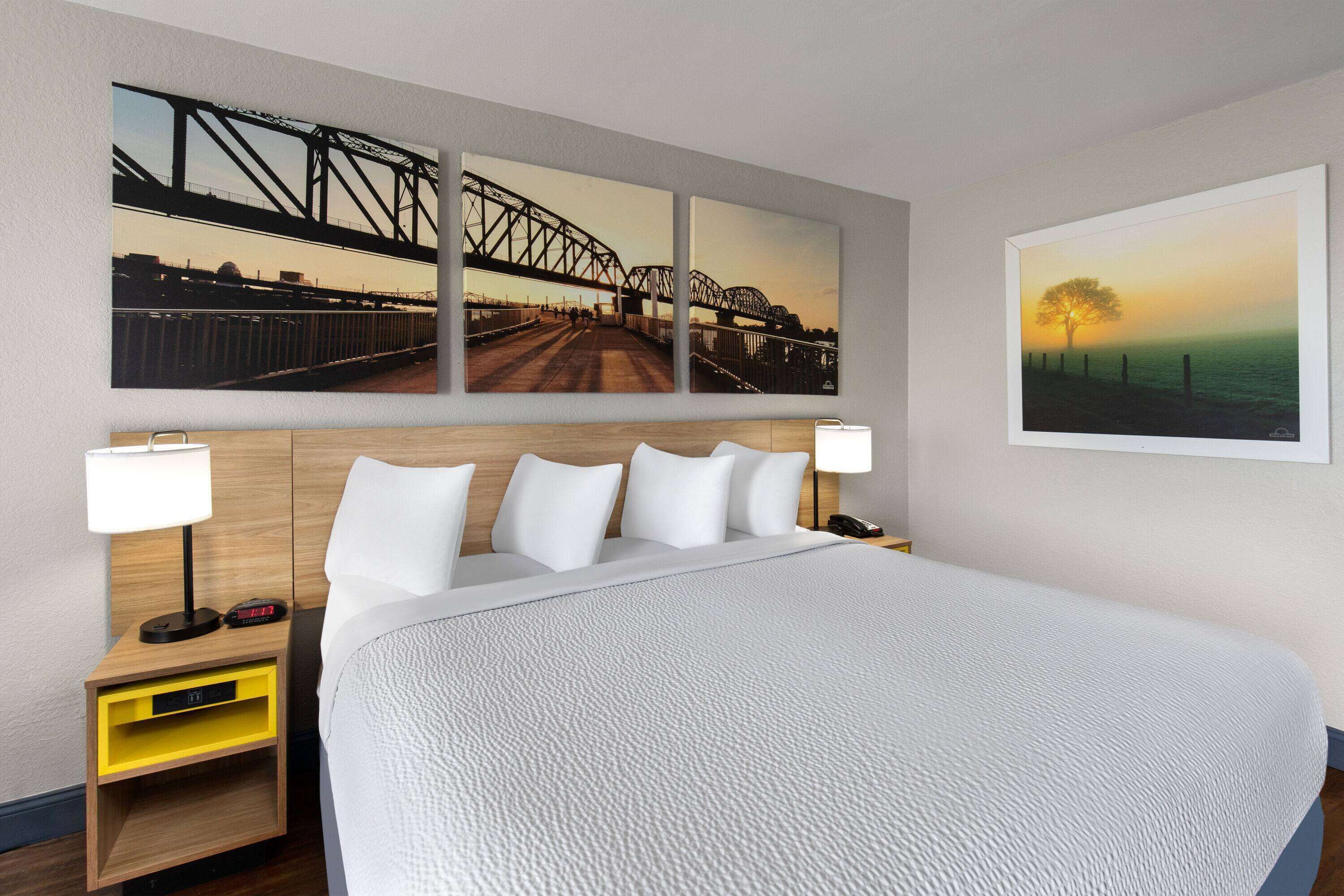 A hotel room with a large bed, two bedside tables with lamps, a nightstand with a clock, and three wall-mounted paintings depicting bridges and a sunset.