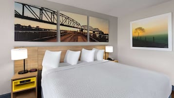 A hotel room with a large bed, two bedside tables with lamps, a nightstand with a clock, and three wall-mounted paintings depicting bridges and a sunset.