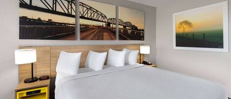 A hotel room with a large bed, two bedside tables with lamps, a nightstand with a clock, and three wall-mounted paintings depicting bridges and a sunset.