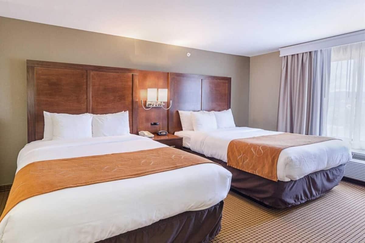 Ein Hotelzimmer mit zwei Betten, einem hölzernen Kopfteil, einem Nachttisch mit Lampe und einem großen Fenster mit Vorhängen.