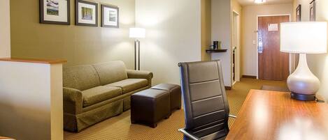 A hotel room with a sofa, a chair, a small table, and a lamp. There are framed pictures on the wall.