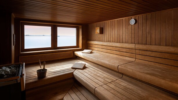Un sauna avec des bancs en bois, une fenêtre donnant sur l’eau, une horloge au mur et une petite table avec un seau et des cuillères en bois.