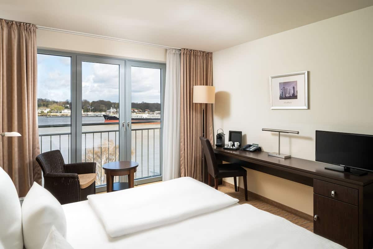 A hotel room with a bed, a desk with a computer, a chair, a TV, and a window with a view of the water and boats.