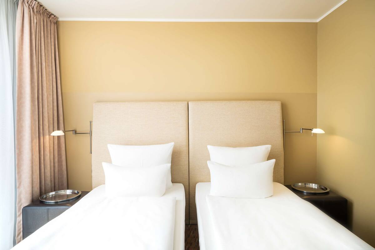 A hotel room with two single beds, a bedside table with a lamp, and a window with curtains.