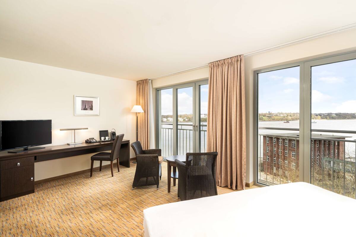A hotel room with a bed, a desk with a computer, a TV, and a view of a body of water through large windows.