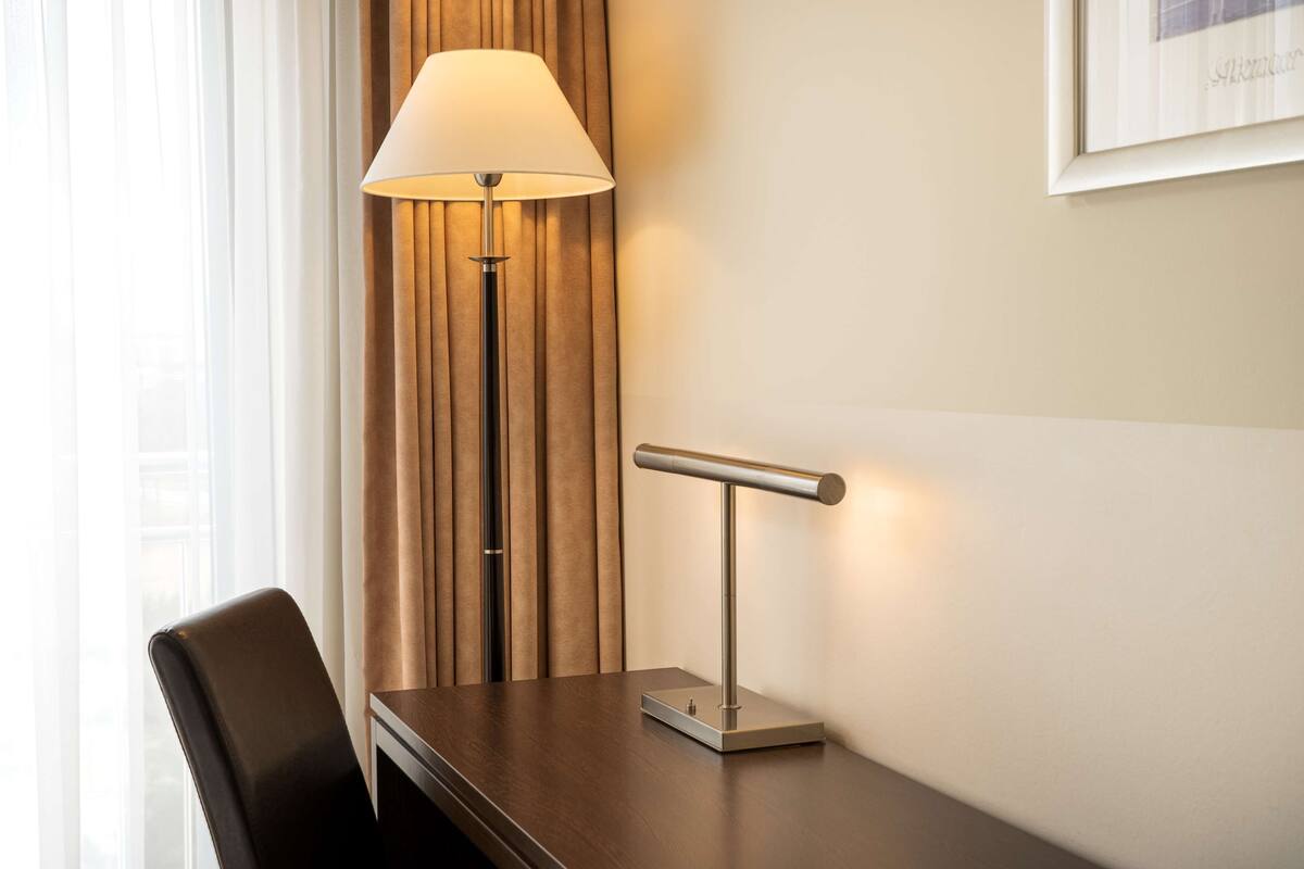 A hotel room with a desk, a lamp, a chair, and a curtain.
