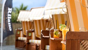 Une rangée de chaises longues en bois, surmontées de parasols à rayures, un verre de jus d’orange et un drapeau noir avec du texte blanc.