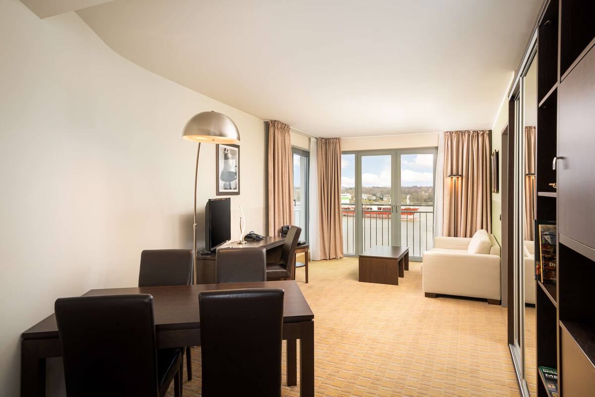 A hotel room with a dining area, a sofa, a TV, and a balcony with curtains.