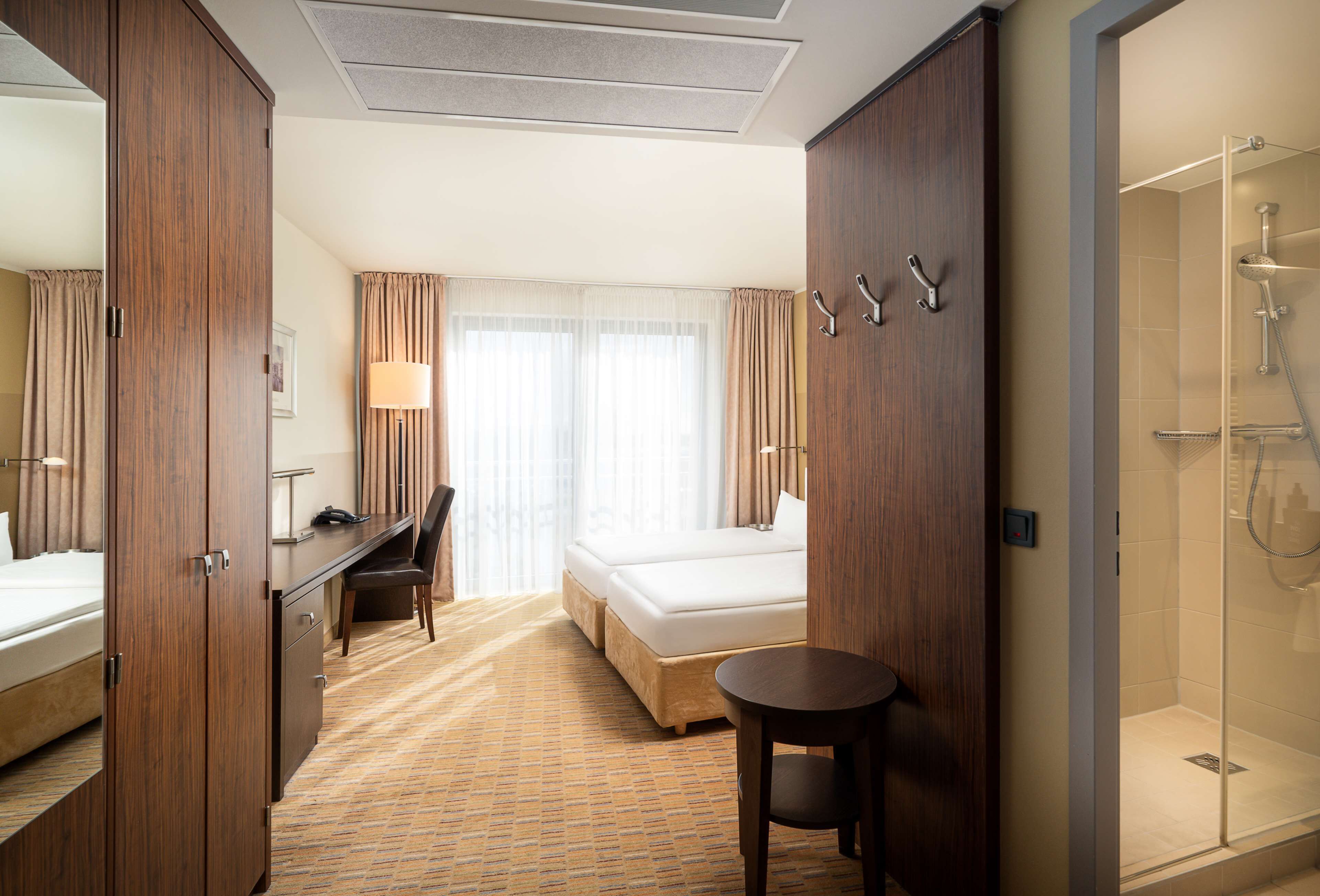A hotel room with a bed, desk, chair, and a small table. There is a shower area visible.