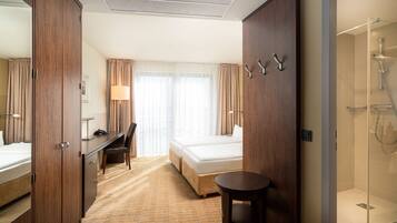 A hotel room with a bed, desk, chair, and a small table. There is a shower area visible.