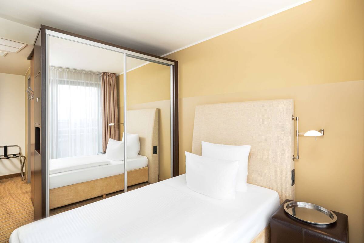 A hotel room with a bed, a bedside table with a tray, a mirror, and a window with curtains.