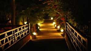 A nighttime view of a bridge with decorative railings and lights leading to a pathway surrounded by foliage. - Hotel Laguna Anjuna, Goa (Anjuna)