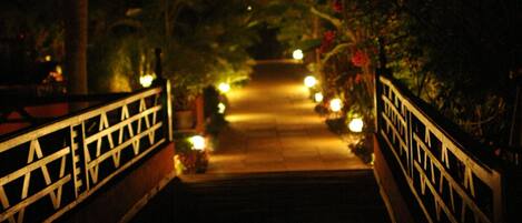 Una vista nocturna de un puente con barandillas decorativas y luces que guían a un sendero rodeado de follaje.