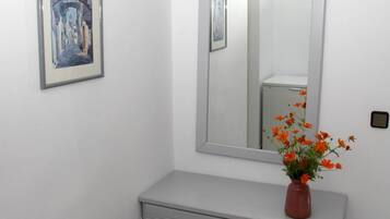 A bathroom with a white vanity, a mirror, a framed picture, a vase with flowers, and an air conditioning unit.