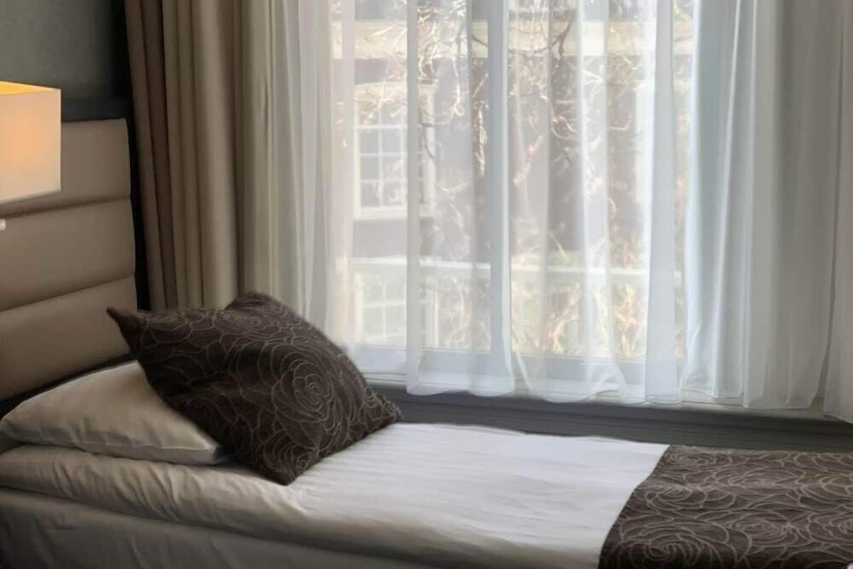 A bed with a patterned pillow and a white bedspread, a bedside lamp, and a window with sheer curtains.