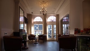 A spacious room with a high ceiling, a chandelier, leather armchairs, a small table, and large windows with a view of buildings outside. - Hotel Clemens Amsterdam (Amsterdam)
