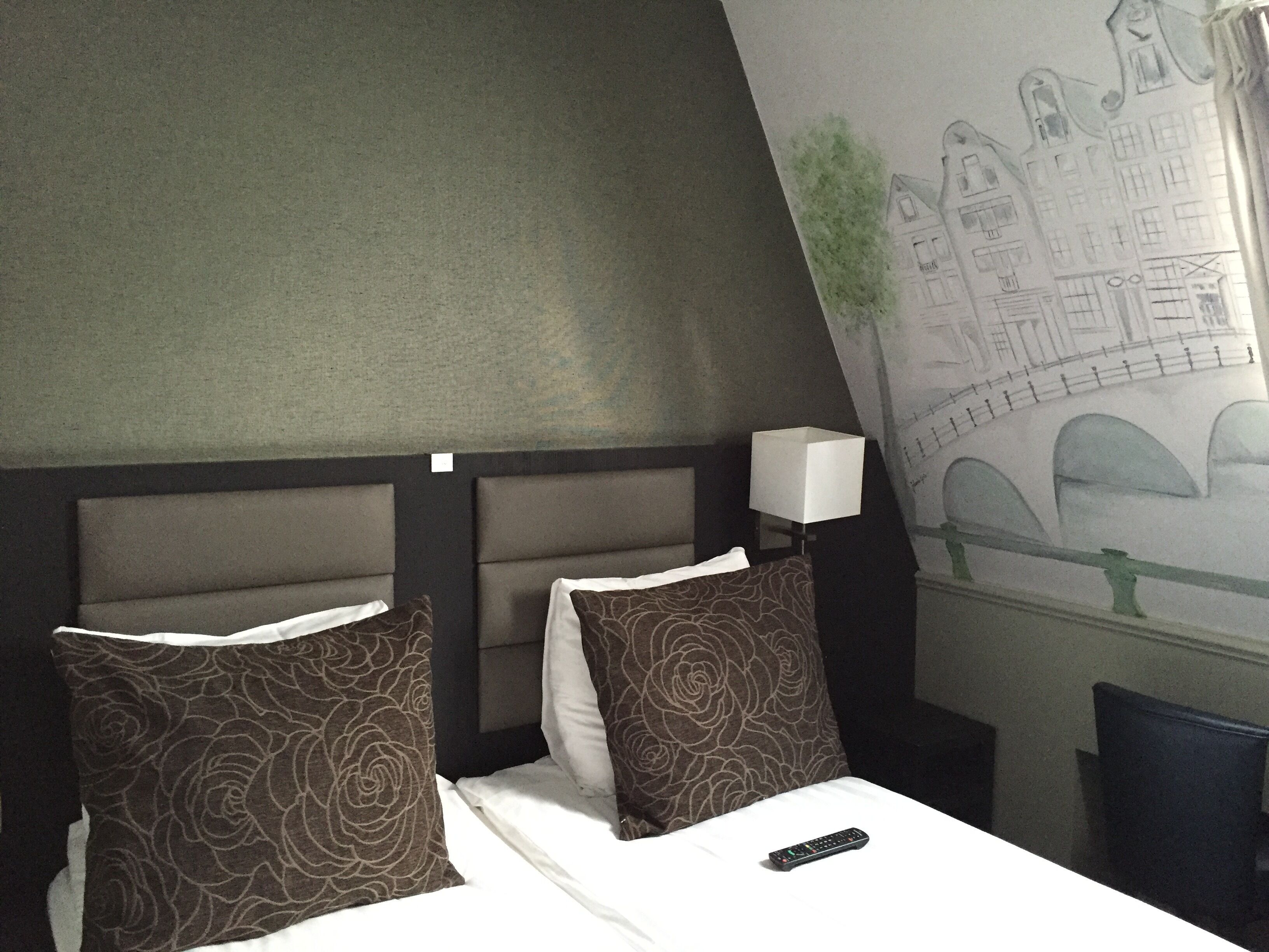 Une chambre d’hôtel avec un lit, deux oreillers, une télécommande et une fresque murale représentant un paysage urbain.