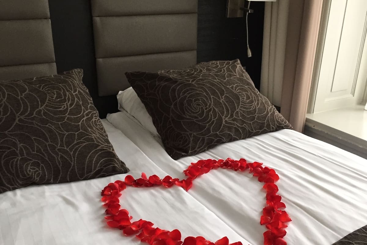 A neatly made bed with a heart-shaped decoration of red rose petals, dark patterned pillows, and a headboard with a lamp.