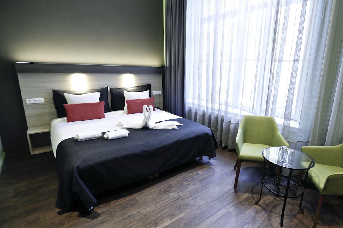 Una habitación de hotel con una cama, un sillón verde, una mesita y una ventana con cortinas.
