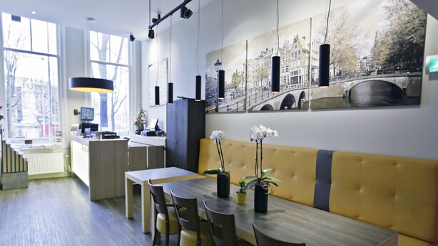 Un comedor moderno con una mesa de madera, sillas y un banco acolchado amarillo. Hay plantas en macetas sobre la mesa y un cuadro de gran formato con una vista urbana en la pared.