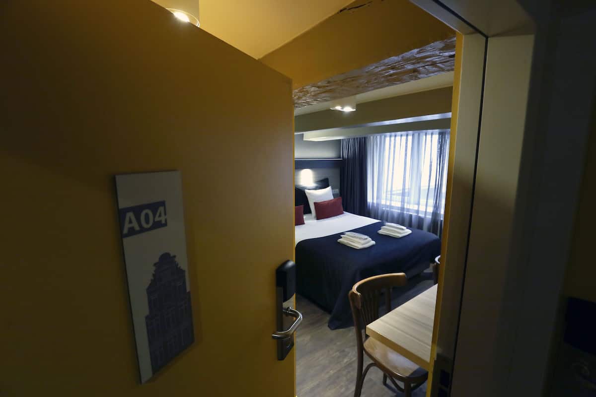 Habitación de hotel con cama, escritorio y silla. Cuenta con una ventana con cortinas, una lámpara de pared y una puerta con manija.