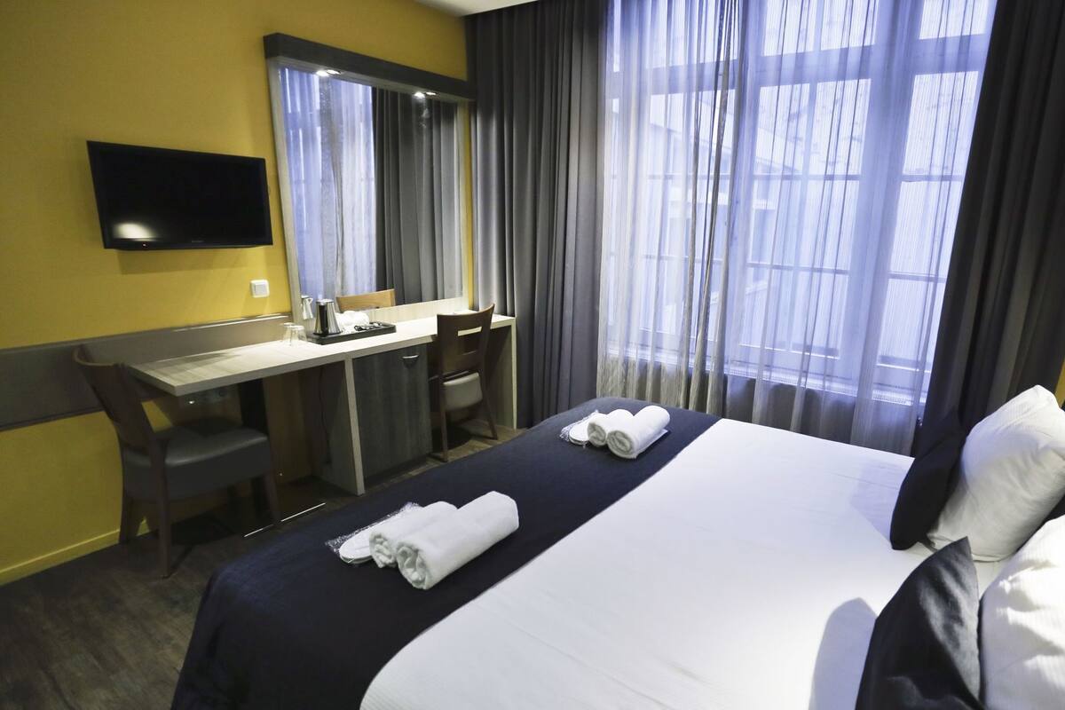 Habitación de hotel con cama, escritorio, silla, televisión de pantalla plana y ventana grande con cortinas.