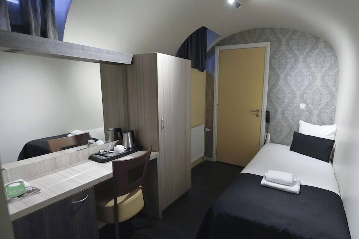 Habitación de hotel pequeña con cama, escritorio con silla, armario y puerta que da a otra habitación.