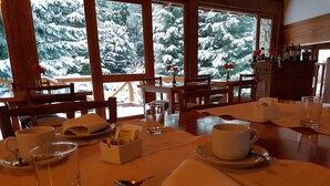 Een eettafel gedekt met borden, bekers en bestek, met uitzicht op besneeuwde bomen door grote ramen.