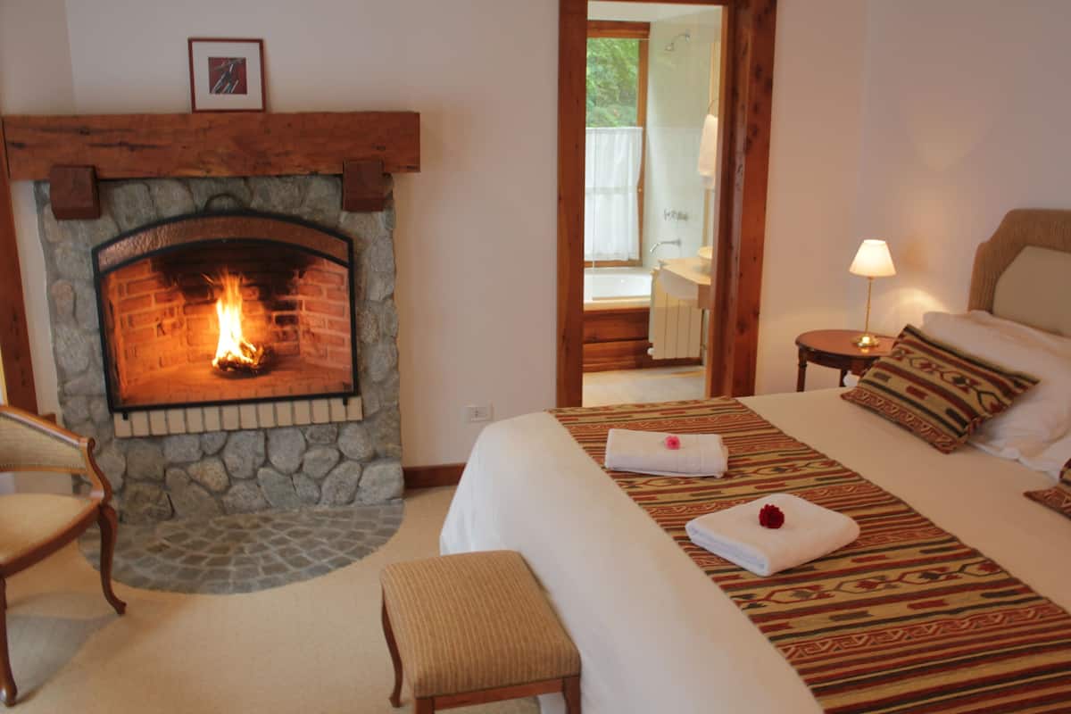 Une chambre avec une cheminée en pierre, un lit recouvert d’une couvre-lit à motifs, une table de chevet avec une lampe, et une vue sur la salle de bain à travers une porte ouverte.