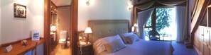 Habitación de hotel con cama, mesita de noche, lámpara y vistas a árboles por la ventana.
