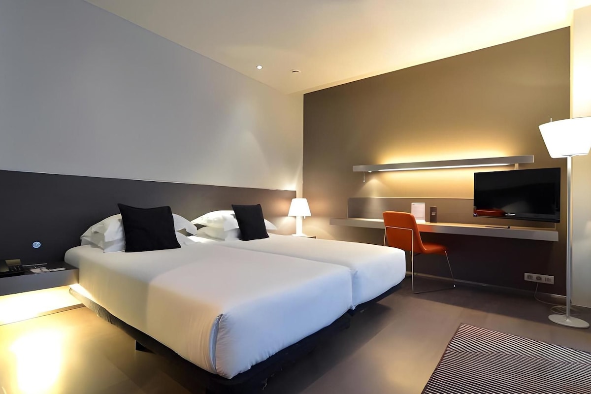 A hotel room with a bed, a desk, a chair, a TV, and a lamp.