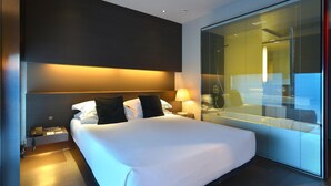 Una habitación de hotel moderna con una cama grande, una mesita de noche con lámpara y un ventanal con persianas.