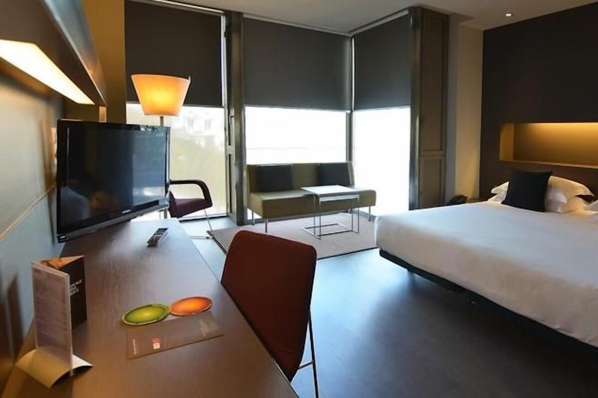 A hotel room with a bed, a desk with a television, a chair, and a window.