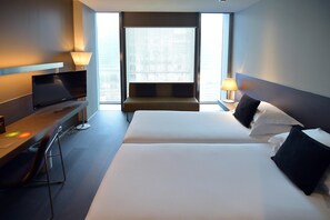 A hotel room with a bed, a desk with a TV, and a window with a view of the city. - Soho Hotel (Barcelona)