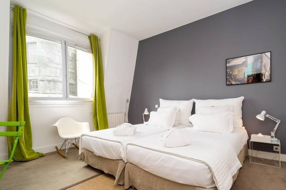 Una cama doble con ropa de cama blanca, una mesita de noche pequeña con una lámpara, una silla y una ventana con cortinas verdes.