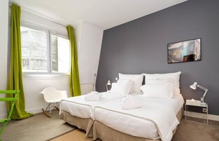 Una cama doble con ropa de cama blanca, una mesita de noche pequeña con una lámpara, una silla y una ventana con cortinas verdes.