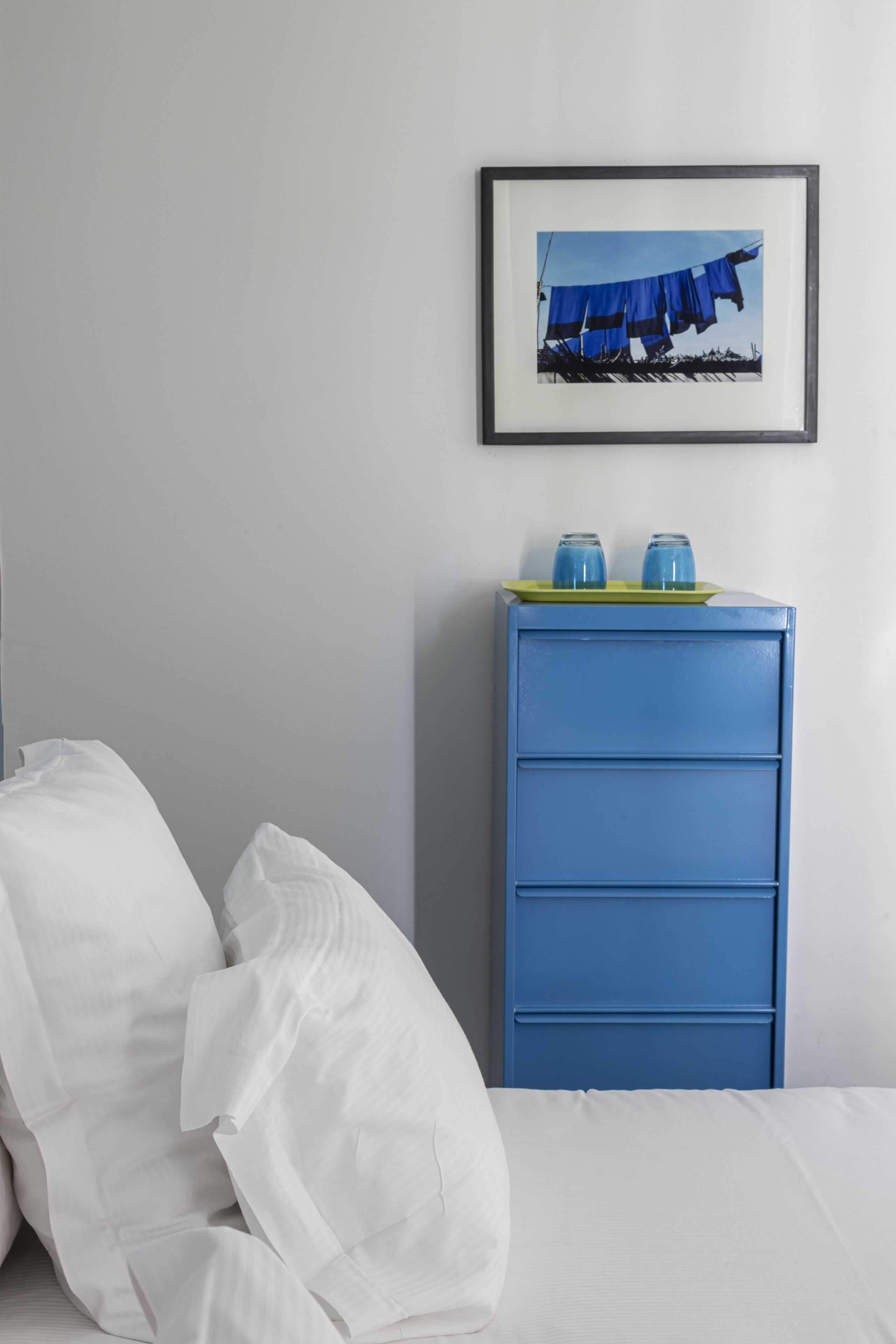 Una cama bien hecha con sábanas blancas, un tocador azul con dos vasos azules encima, y un cuadro colgado en la pared que muestra un paraguas azul.