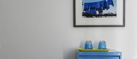 Una cama bien hecha con sábanas blancas, un tocador azul con dos vasos azules encima, y un cuadro colgado en la pared que muestra un paraguas azul.