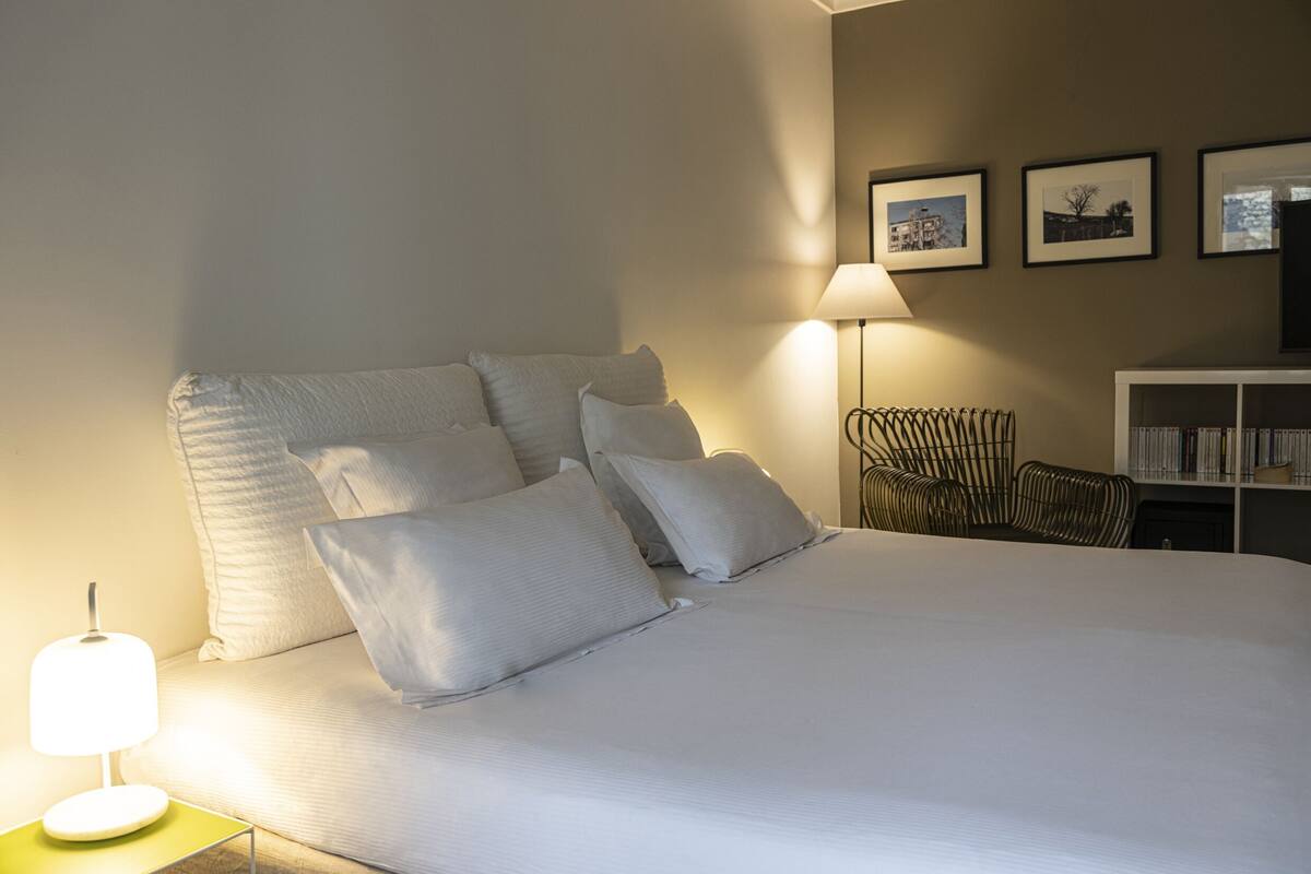 Una cama bien hecha con almohadas, una lámpara de noche, una silla y una estantería en una habitación de hotel.