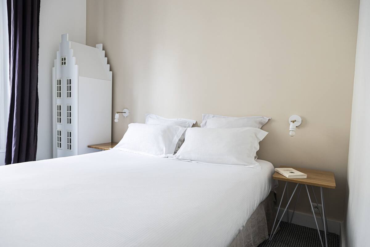 Una cama bien tendida con sábanas blancas, una mesita de noche con un libro y una lámpara moderna fijada a la pared.